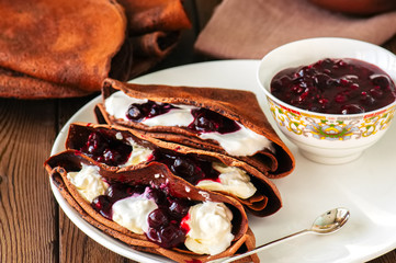 Chocolate crepes (blinis) with sour cream and black currant sauce on a white plate. Wooden...