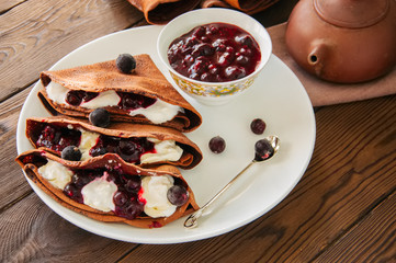 Chocolate crepes (blinis) with sour cream and black currant sauce on a white plate. Wooden background.