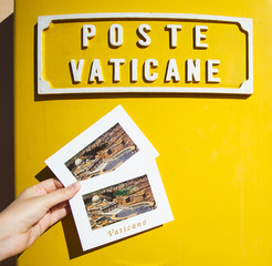 Hand holding two postcards with yellow vatican mailbox in the background. St. Peter's Basilica,...