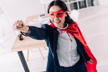 attractive super businesswoman in cape and mask standing with hand up in office