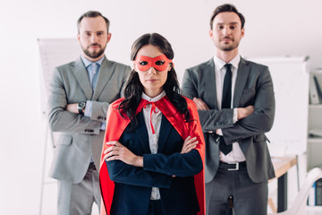 super businesswoman standing with crossed arms in mask and cape with businessmen in office