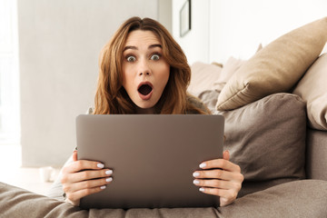 Image of excited european woman lying on sofa in living room, and expressing interest or surprise while using laptop