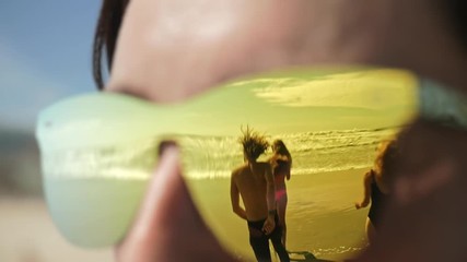 Reflection in sunglasses of friends running to sea. Close up of girl with glasses reflected young happy people enjoying summer vacation on beach together. Travel, dream, memories, youth concept.