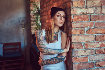 A tattoed sexy blonde in a t-shirt and a hat posing against a brick wall.