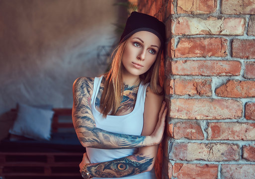 A tattoed sexy blonde in a t-shirt and a hat posing against a brick wall.