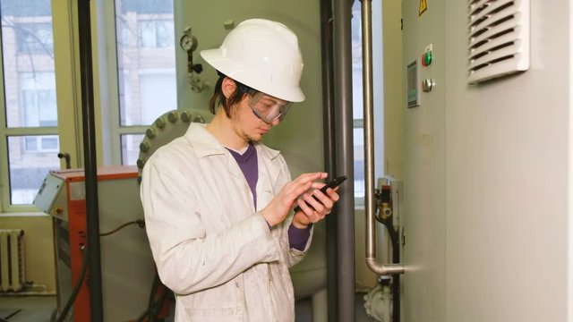 The engineer in a white helmet and goggles writes data to the smartphone from the display of the electrical control cabinet, uses the touch screen tablet to write data.