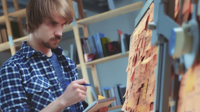 Creative man in a blue checkered shirt is standing in front of a board with orange stickers, writing ideas. Brainstorming, making mind map. Creative, young adult man in relaxed working mode.