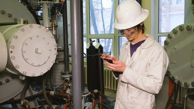 The technologist in a protective helmet and glasses makes inspection of the chemical installation, enters data into the smartphone, uses a touch-screen tablet to record data.