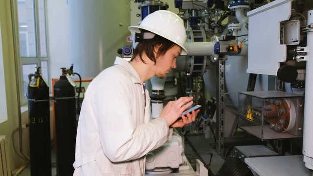 Engineer technologist using a smartphone with a touch screen performs inspection and data acquisition from a gas installation, in a white protective helmet and dressing gown.