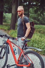 Fototapeta na wymiar A handsome redhead male with a stylish haircut and beard dressed in sportswear and sunglasses walks in the park with a bicycle and backpack.