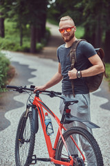 A handsome redhead male with a stylish haircut and beard dressed in sportswear and sunglasses walks in the park with a bicycle and backpack.