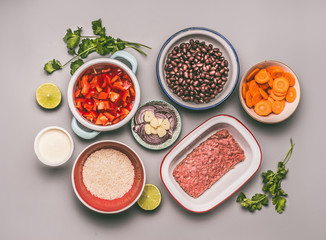 Obraz na płótnie Canvas Flat lay of bowls with cooking ingredients for balanced one pan meal with beans, minced meat, rice and various cut vegetables on gray background, top view