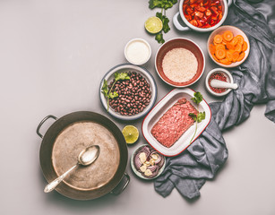 Bowls with ingredients for balanced one pan meal with beans, minced meat, rice and various cut vegetables and cooking pot with spoon on gray background, top view