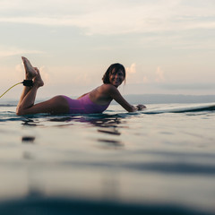 smiling girl lying on surfboard in water in ocean at sunset