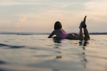 young woman lying on surfboard in water and looking at sunset