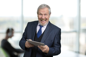 Extremely happy man in suit with tablet. Portrait of rejoicing old manager in office. Bright windows background.