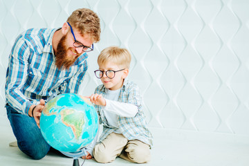 Happy beard dad and son sitting on the floor and looking at globe together at home with white polygonal background.