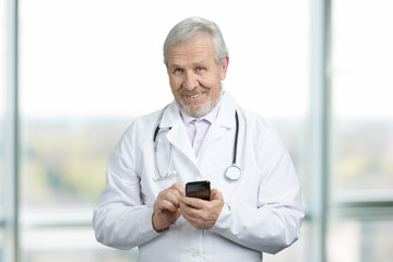 Old doctor standing, holding and typing on mobile phone. Smiling senior physician with phone looking at camera.