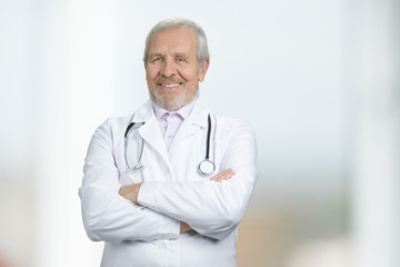 Portrait of doctor standing with arms crossed in hospital. Wrinkled caucasian physician. Bright blurred background.
