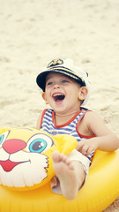 Little Caucasian boy in sailor suit and cap sitting on the beach in swim ring and laughs.