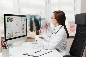 Female doctor sitting at desk, working on computer, holding X-ray of lungs, fluorography, roentgen in light office in hospital. Woman in medical gown, stethoscope in consulting room. Medicine concept.