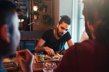 Multiracial friends eat breakfast in lounge bar. Young men chat while having tasty food and drinks.
