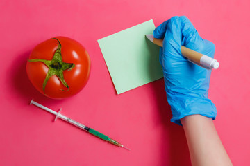GMO Scientist Make Note, Green Liquid in Syringe,Red Tomato - Genetically Modified Food Concept on Pink Background.