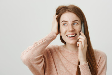 Indoor portrait of feminine european ginger girl with freckles talking on phone while scratching...