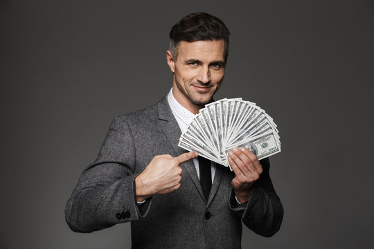 Photo of happy man 30s in formal costume demonstrating lots of money dollar banknotes and pointing finger on bills, isolated over gray background