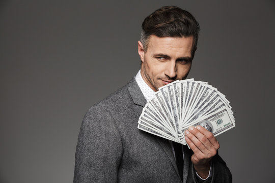 Photo of lucky guy 30s in business suit demonstrating money prize in dollar bills and looking on camera, isolated over gray background