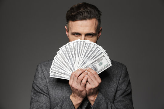 Photo of confident guy 30s in business suit holding fan of money dollar bills at face and looking on camera with strict gaze, isolated over gray background