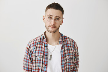 Portrait of sad gloomy guy with glasses, wearing plaid shirt over white t-shirt, sucking lips and frowning, hesitating over gray background. Man wants to say truth but knows it will upset girlfriend