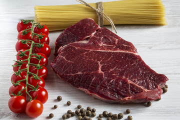 Raw meat, beef steak, brench of ripe cherry tomatoes, spaghetti, health food, top view. White wooden background.
