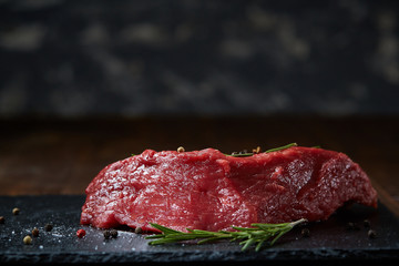 Raw meat beef steaks on black slate board with spices and rosemary over wooden background, copy...