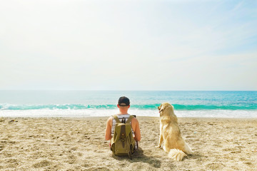 Rear view of man & his dog sitting at beach watching ocean waves, clear sunny day. Fit programmer freelancer male working on laptop computer at sea w shepherd canine. Background, copy space, landscape