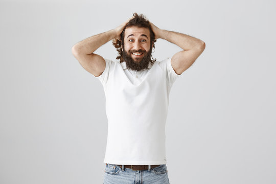 Studio Shot Of Mature Eastern Bearded Male With Broad Smile Coming Hair With Hands On Head, Standing In White Shirt Over Gray Background, Being Amazed And Happy To Receive Great News Or Offer