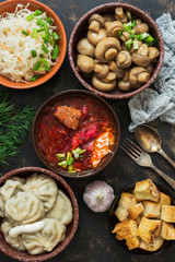 Borsch, dumplings, sauerkraut, pickled mushrooms. Russian dishes on a dark rustic background, top view. Traditional Russian cuisine.