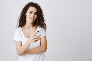 Girl checking messages while ordering lunch in cafe. Good-looking young student with curly hair in casual white t-shirt holding smartphone, smiling at camera, staying online with new device