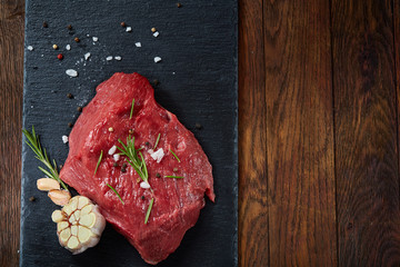 Raw meat beef steaks on black slate board with spices, garlic and rosemary over wooden background, selective focus