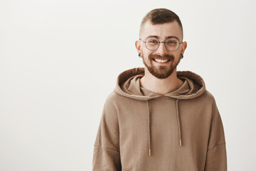 Waist-up indoor shot of friendly handsome hipster guy with trendy haircut and glasses smiling cheerfully while standing against gray background, talking casually with coworkers, being in good mood