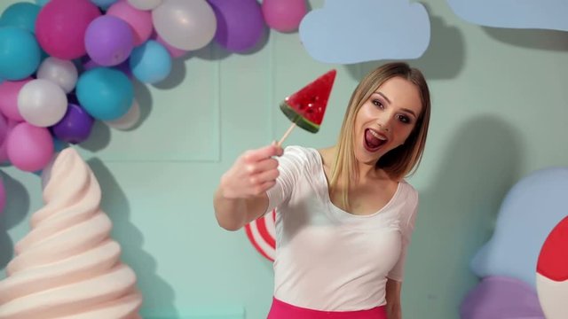 Portrait of a happy girl with a big candy on a background of multi-colored balls, she looks at the camera and offers a candy to you. Barbie. Lollipop.