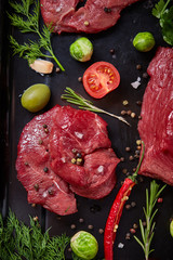 Flat lay of raw beefsteak with vegetables, herbs and spicies on metal tray, close-up, selective focus
