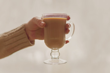 Hot coffee with milk in a transparent glass in female hands, toned