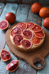 Tasty homemade cake with blood orange slices served with raw blood oranges on oak cutting board over shabby rustic blue background