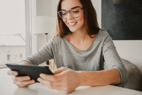 Portrait Of Young Caucasian Woman Wearing Glasses Sitting At Table At Home, Using Digital Tablet, And Smiling