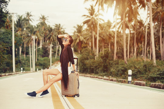 Beautiful style girl seating with suitcase on tropical road