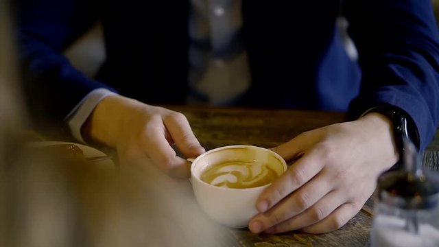 young man is wearing dark blue suit is drinking coffee in restaurant, sitting at table with blonde