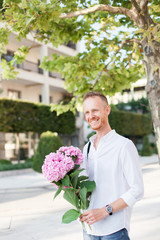 Fototapeta premium Man with smile is waiting for woman with bouquet of pink hydrangea flowers. Guy is wearing in casual white shirt in green city park outdoor at dating.