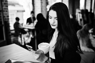 Brunette girl sitting on cafe with cup of cappuccino, working with red laptop and looking at mobile phone.