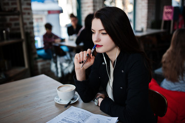 Brunette girl sitting on cafe with cup of cappuccino, listening music on headphones and write some documents.